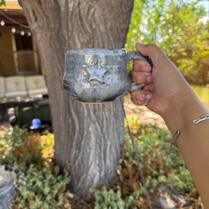 Könnte beinhalten: Eine handgefertigte Keramiktasse mit blauem Glanz und Sternmuster. Die Tasse hat einen Henkel und ein rustikales, strukturiertes Aussehen. Die Tasse ist für Heißgetränke konzipiert.
