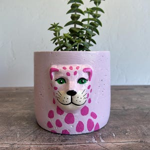 May include: A lavender-coloured planter pot featuring a sculpted leopard face and pink spots. The leopard has green eyes and a white muzzle. The pot contains a green succulent plant. The planter is made of a textured material.