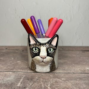 May include: A ceramic cat-shaped pen holder with a black and white face and green eyes. The holder is filled with colorful markers in shades of red, orange, yellow, purple, and pink. The pen holder is on a wooden surface.