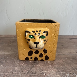Puede incluir: Maceta cuadrada de cerámica de color beige con una cara de leopardo esculpida. El leopardo tiene ojos verdes, manchas negras y una nariz negra. La superficie de la maceta tiene un aspecto texturizado.