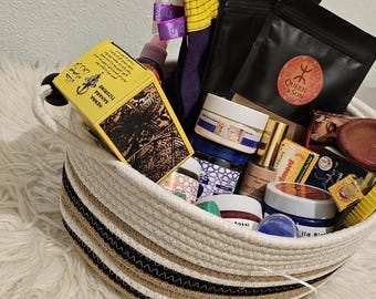 May include: A woven rope basket with white, tan, and black stripes containing various skincare and cosmetic products. Items include a yellow box labeled Henna Sahara Tazarine, black pouches with a Queen & Son logo, jars with blue and white lids, and a yellow comb.