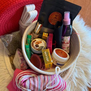 Op de afbeelding: Een gevlochten mand gevuld met schoonheidsproducten, waaronder een rode hoed, een roze en wit gestreepte sjaal en diverse potten en flessen. De mand bevat items met het "Queen & Son" logo, een kleine roze borstel en een zwart zakje.