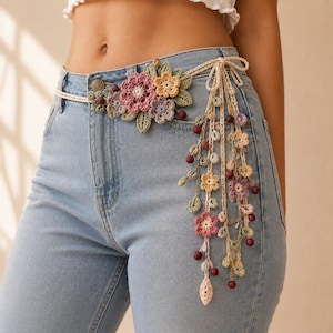 May include: A light blue denim belt featuring a decorative crochet floral arrangement in shades of pink, yellow, and green. The belt includes dangling crochet flower and leaf motifs accented with small dark red beads, tied in a bow at the side.