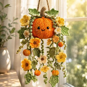 May include: A hanging crochet decoration featuring a central orange pumpkin with a smiling face, surrounded by green leaves, yellow and white flowers, and smaller orange pumpkins. The entire arrangement is suspended by a thick tan rope cord.