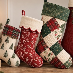 May include: Three Christmas stockings featuring varied designs. The left stocking is cream with green pine trees and a plaid cuff. The middle stocking is red with white sprigs and a scalloped cream cuff. The right stocking has a quilted patchwork pattern.