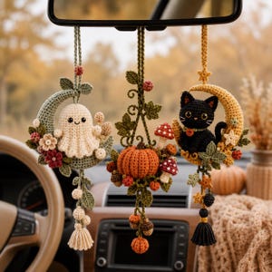 Könnte beinhalten: Drei hängende Häkelornamente für den Rückspiegel im Auto. Eines zeigt ein cremefarbenes Gespenst auf einem salbeigrünen Halbmond, eines einen orangefarbenen Kürbis mit Pilzen und Ranken, und eines eine schwarze Katze auf einem gelben Halbmond.