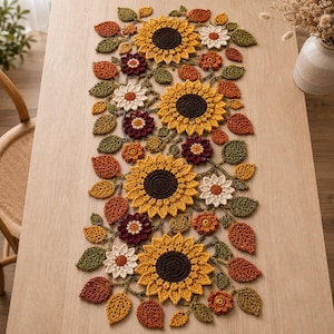 May include: A rectangular crochet table runner featuring a repeating pattern of large yellow sunflowers with dark brown centers, smaller white and maroon flowers, and various autumn-colored leaves in shades of orange, rust, and olive green.