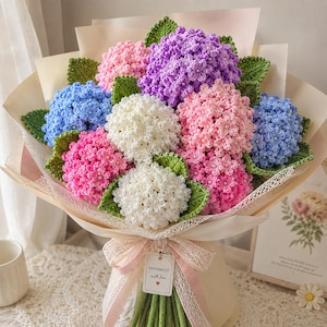 May include: A bouquet of crocheted hydrangea flowers in shades of purple, pink, blue, and white, accented with green crocheted leaves. The stems are wrapped in light-colored paper and lace ribbon with a tag reading HANDMADE with love.