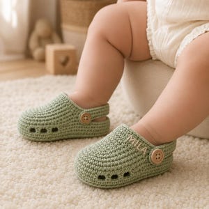 May include: A pair of sage green crocheted baby clogs featuring a rounded toe, three ventilation holes on the side, and a pivoting heel strap secured with a small wooden button. The right shoe is embroidered with the name MILA in white thread.