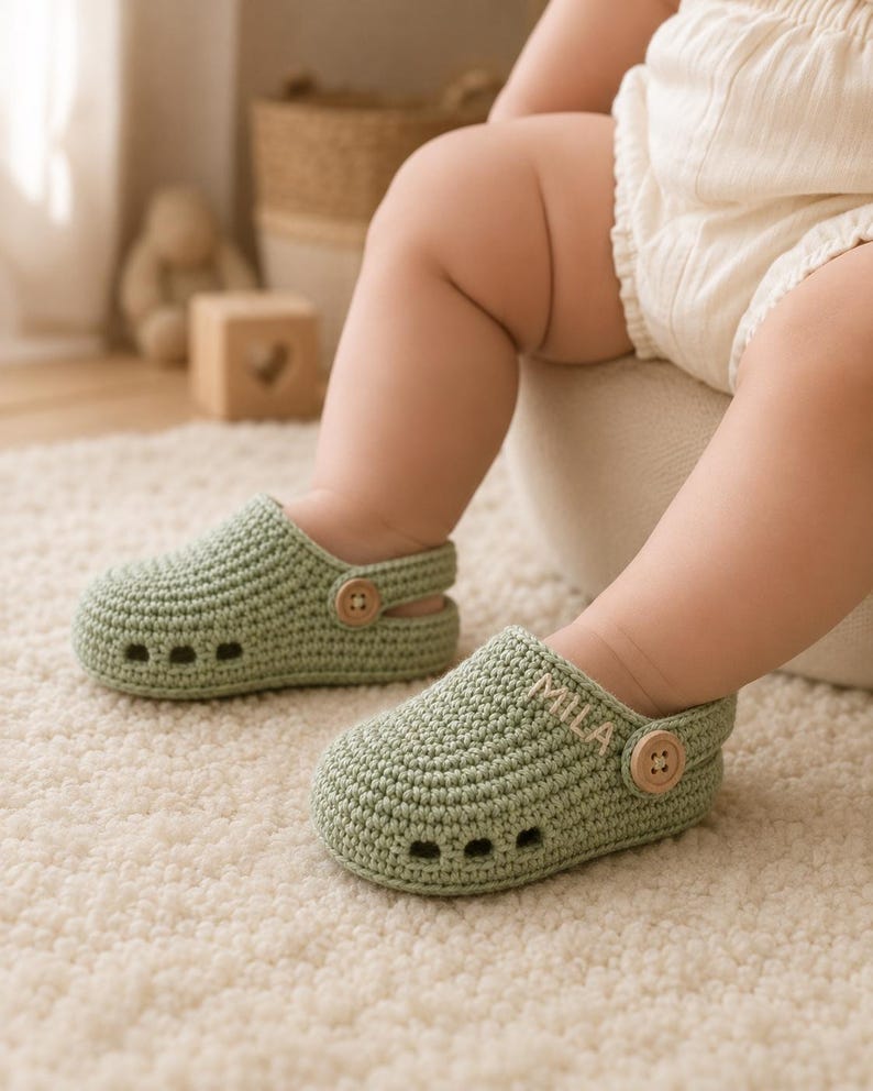 May include: A pair of sage green crocheted baby clogs featuring a rounded toe, three ventilation holes on the side, and a pivoting heel strap secured with a small wooden button. The right shoe is embroidered with the name MILA in white thread.
