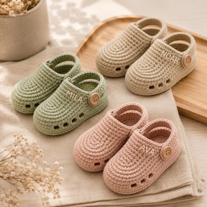 Op de afbeelding: Drie paar handgemaakte gehaakte babyklompjes in saliegroen, beige en oudroze. Elk paar heeft een ronde neus, zijdelingse uitsparingen, een bandje met een houten knoop en de namen Mila, Noah en Ava in de zijkant geborduurd.