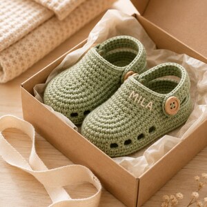 May include: A pair of sage green crocheted baby shoes with a rounded toe, side cutouts, and a wooden button strap. The name MILA is embroidered in white thread on the side of one shoe. The shoes are presented inside a brown cardboard gift box.