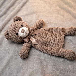 May include: A handmade brown crochet teddy bear security blanket with a white muzzle and a brown satin ribbon. The bear has black eyes and a textured, plush appearance. The blanket is lying on a gray, textured surface.