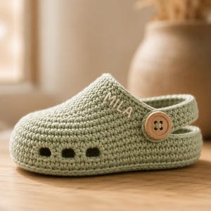 May include: A sage green crocheted clog featuring a rounded toe, three small ventilation holes at the front, and a movable heel strap secured by a light-coloured wooden button. The name MILA is embroidered in white thread on the side.