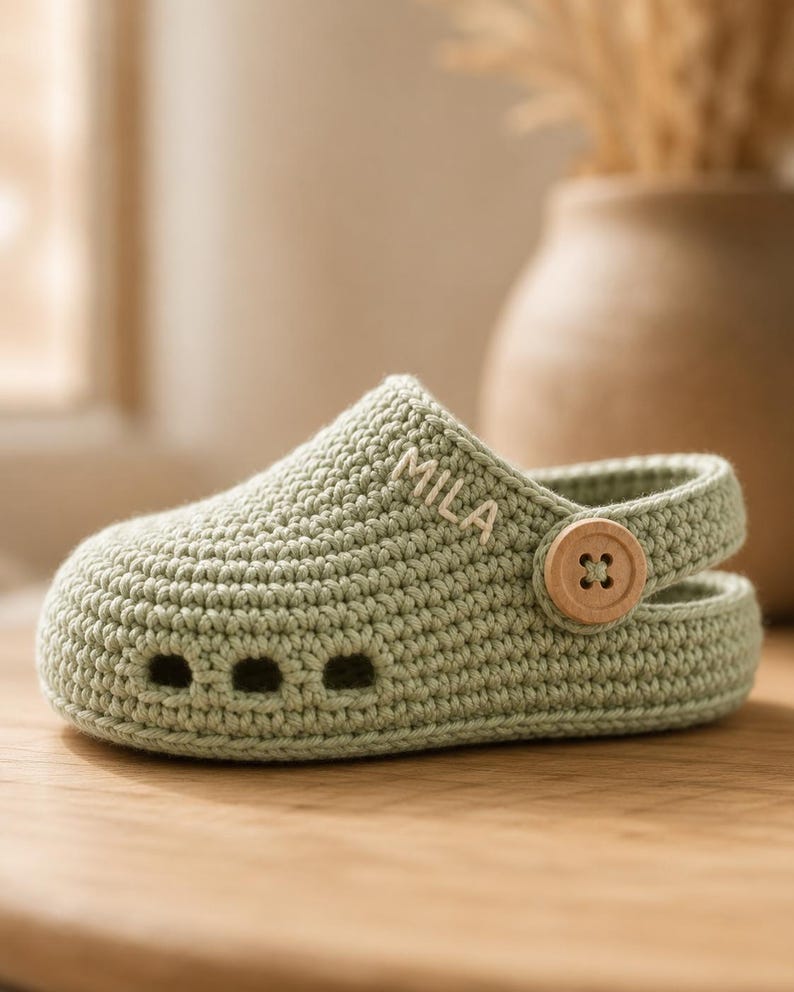 May include: A sage green crocheted clog featuring a rounded toe, three small ventilation holes at the front, and a movable heel strap secured by a light-coloured wooden button. The name MILA is embroidered in white thread on the side.