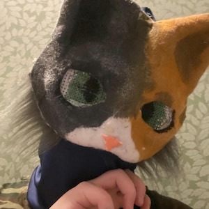 May include: A handmade cat mask with a calico pattern. The mask features black, orange, and white fur, with green mesh eyes and a small orange nose. The mask is worn over the head, with a dark blue fabric at the neck.