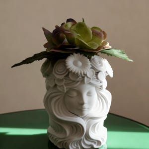 May include: A white ceramic planter shaped like a human head with closed eyes and flowing hair. The top of the head is adorned with sculpted floral details, and a large, artificial green and purple flower is placed inside the opening.