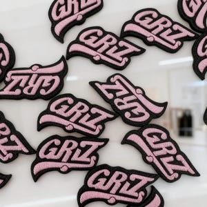 Puede incluir: Una colección de parches bordados con el texto "GRIZ" en una fuente estilizada y negrita. Cada parche tiene un interior rosa claro con un borde negro grueso, creando un diseño de alto contraste. Los parches están dispersos por el encuadre.