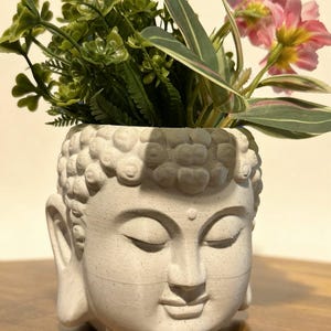 Puede incluir: Una maceta blanca con forma de cabeza de Buda llena de plantas artificiales. La maceta tiene una cara serena con los ojos cerrados. Las plantas incluyen follaje verde y flores rosas. La maceta está sobre una superficie de madera.