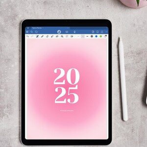 Może przedstawiać: Cyfrowy tablet wyświetlający różowe gradientowe tło z białymi cyframi "2025". Biały rysik spoczywa obok tabletu. Tekst "Taylor's Planner" jest widoczny u góry ekranu.