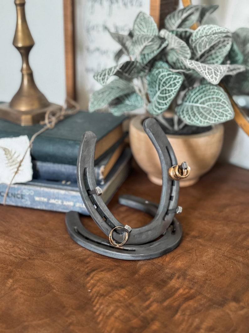 May include: A rustic horseshoe ring holder made of dark gray metal. Two horseshoes are welded together, with small metal pegs to hold rings. Two gold rings are displayed on the holder, which sits on a wooden surface.
