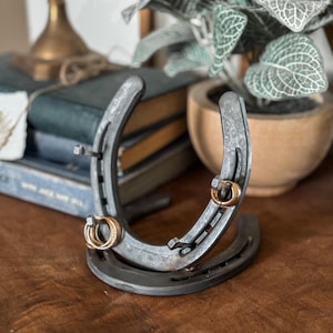 May include: A rustic metal horseshoe ring holder with two gold rings. The horseshoe is a dark gray color and sits on a wooden surface. A stack of books and a potted plant are in the background.