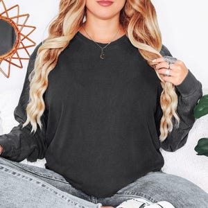 May include: A charcoal grey long-sleeved sweatshirt. The sweatshirt is plain and has a crew neck. The model is wearing the sweatshirt with light wash jeans. The model is also wearing a necklace and rings.
