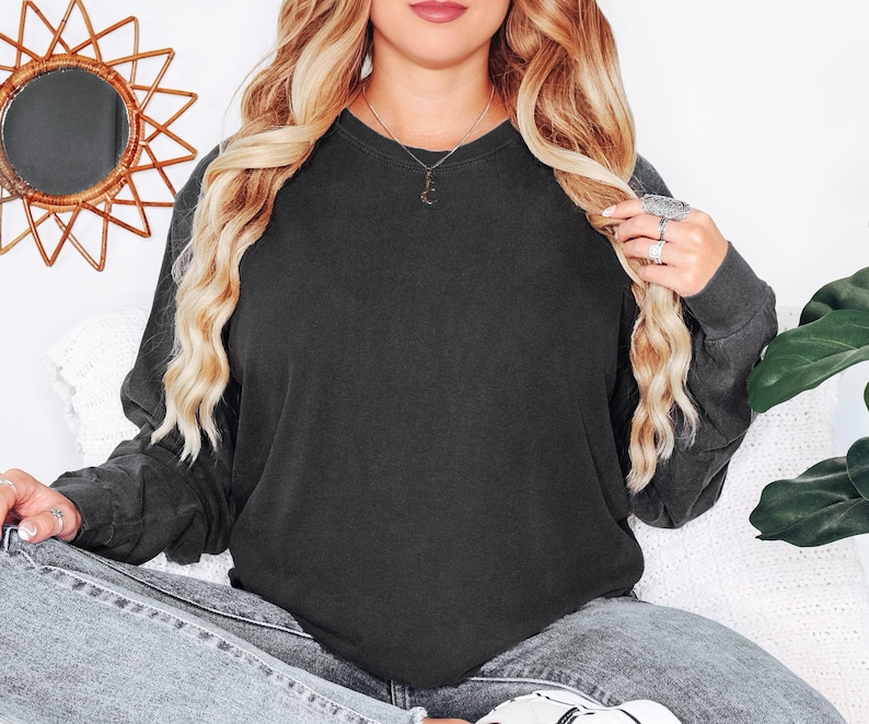 May include: A charcoal grey long-sleeved sweatshirt. The sweatshirt is plain and has a crew neck. The model is wearing the sweatshirt with light wash jeans. The model is also wearing a necklace and rings.