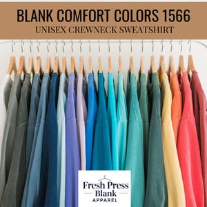 May include: A selection of unisex crewneck sweatshirts in a spectrum of colours, displayed on wooden hangers. The sweatshirts vary from grey to red hues. The image includes the text "BLANK COMFORT COLORS 1566" and "UNISEX CREWNECK SWEATSHIRT". The Fresh Press Blank Apparel logo is visible.