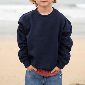 May include: A navy blue crew neck sweatshirt. The sweatshirt is worn over a red top and paired with blue jeans. The person is standing on a sandy beach with the sea in the background. The sweatshirt is a solid colour.