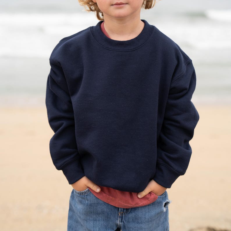May include: A navy blue crew neck sweatshirt. The sweatshirt is worn over a red top and paired with blue jeans. The person is standing on a sandy beach with the sea in the background. The sweatshirt is a solid colour.