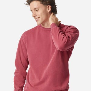 May include: A crew neck sweatshirt in a muted red colour. The long-sleeved top has a ribbed hem and cuffs. The model is wearing the sweatshirt and is smiling.