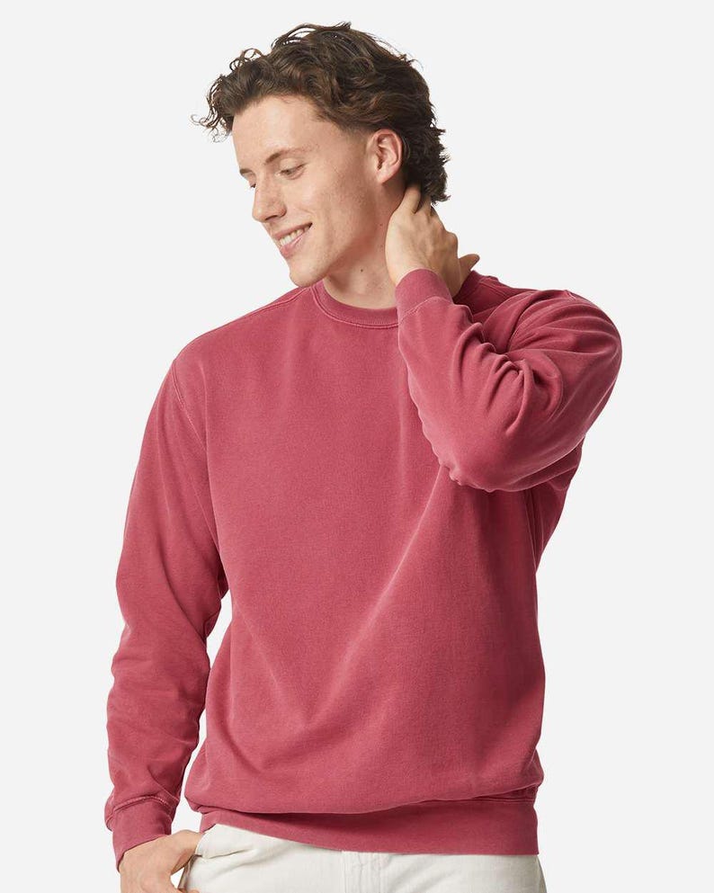 May include: A crew neck sweatshirt in a muted red colour. The long-sleeved top has a ribbed hem and cuffs. The model is wearing the sweatshirt and is smiling.