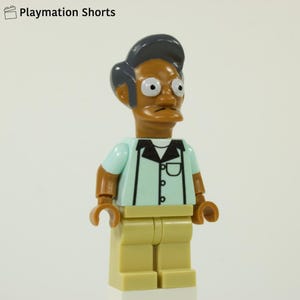 Puede incluir: Una figura de Lego de un personaje con piel oscura, peinado negro y ojos grandes y expresivos. La figura lleva una camisa azul claro con cuello negro y pantalones color beige. La figura es un personaje de Playmation Shorts.