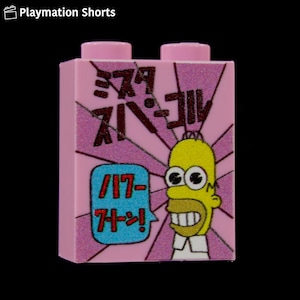 May include: A pink building block featuring a cartoon image of Homer Simpson with Japanese text. The block has a pink background with a sunburst design and a speech bubble with more Japanese text. The top of the block has two pink studs.