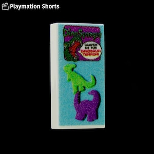 May include: A rectangular, white-edged block with a light blue surface. The block features a purple graphic with the text "Dino Sponge" and a green dinosaur illustration. Below, there are two dinosaur illustrations, one green and one purple. The text "Dampen Me For Dinosaur Terror" is also visible.