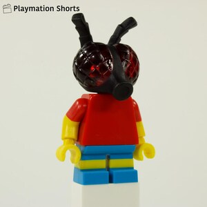 Może przedstawiać: Minifigurka Lego z czerwonym tułowiem, żółtymi ramionami i niebiesko-żółtymi nogami. Figurka ma czarną maskę i anteny oraz czerwoną, przezroczystą głowę. Obraz zawiera tekst "Playmation Shorts" w lewym górnym rogu.