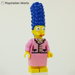 Puede incluir: Una figura de Lego de Marge Simpson, con su icónico pelo azul alto, piel amarilla y un traje rosa con falda a juego. La figura tiene detalles negros y un collar amarillo. La figura está sobre una base amarilla.