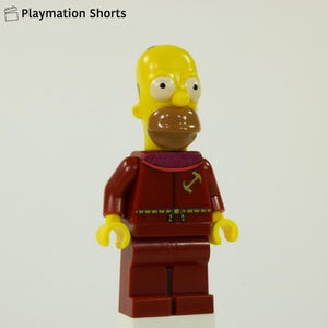 May include: A Lego figure of Homer Simpson in a red uniform with a yellow head and arms. The figure has a brown mustache and a purple collar. The text "Playmation Shorts" is in the top left corner.