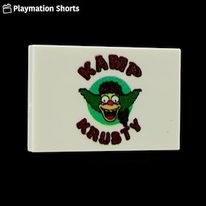 May include: A rectangular, off-white block featuring a cartoon image of Krusty the Clown from The Simpsons. The image includes the words "KAMP KRAUSTY" in a glittery, maroon font, with Krusty's face in the center.