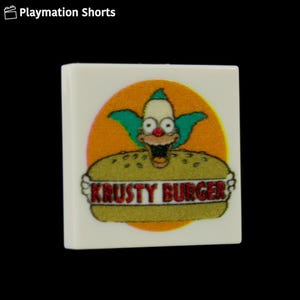 May include: A white square featuring a cartoon image of Krusty the Clown over a Krusty Burger. The clown has green hair, a red nose, and a wide grin. The background is orange.