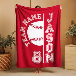 May include: A red baseball-themed blanket with white text and graphics. The design includes a baseball, the name "JASON", the number "8", and the words "TEAM NAME". The blanket is made of a soft material.