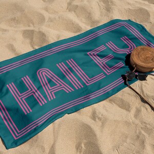 Puede incluir: Toalla de playa turquesa con el nombre "HAILEY" en fuente retro rosa. La toalla tiene un borde rosa y est&aacute; sobre arena. Una bolsa redonda tejida y gafas de sol est&aacute;n al lado de la toalla.