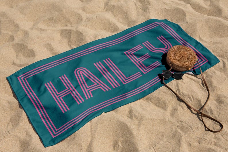 Puede incluir: Toalla de playa turquesa con el nombre "HAILEY" en fuente retro rosa. La toalla tiene un borde rosa y est&aacute; sobre arena. Una bolsa redonda tejida y gafas de sol est&aacute;n al lado de la toalla.