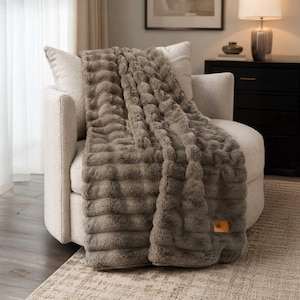 May include: A taupe-colored faux fur throw blanket with a textured, ribbed, and tufted surface pattern. The blanket features a small, rectangular, light brown leather-like patch detail near the bottom corner, adding a subtle accent.