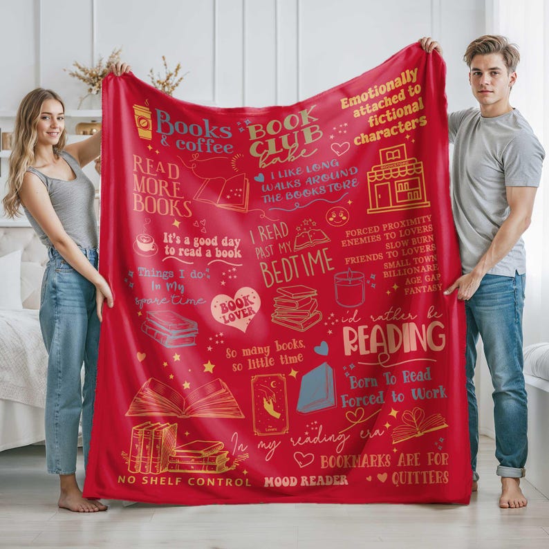 May include: A red fleece blanket with gold and white text and illustrations related to reading. The blanket features phrases like "Books & coffee," "Book Club babe," and "I'd rather be reading." It is held by two people.