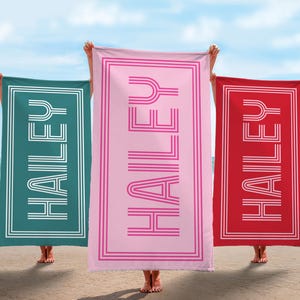 Puede incluir: Tres toallas de playa rectangulares, una turquesa, una rosa y una roja, con el nombre "HAILEY" en blanco en una fuente retro. Las toallas tienen un borde blanco y se sostienen en una playa con el mar de fondo.