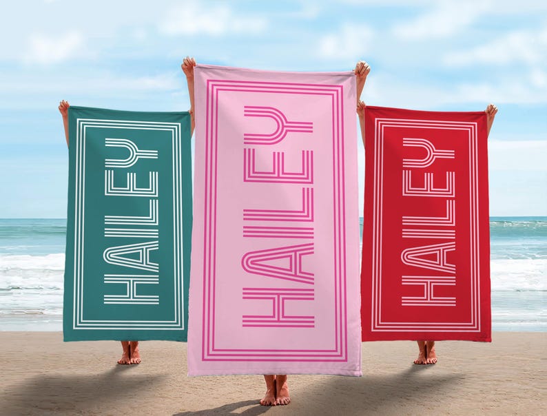 Puede incluir: Tres toallas de playa rectangulares, una turquesa, una rosa y una roja, con el nombre "HAILEY" en blanco en una fuente retro. Las toallas tienen un borde blanco y se sostienen en una playa con el mar de fondo.