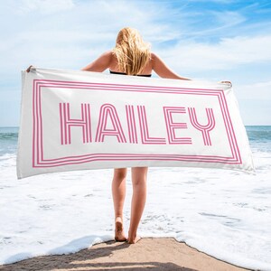Puede incluir: Una toalla de playa blanca con el nombre "HAILEY" en rosa, enmarcada por l&iacute;neas rosas. La toalla es sostenida por una persona en una playa con el oc&eacute;ano y el cielo azul de fondo.