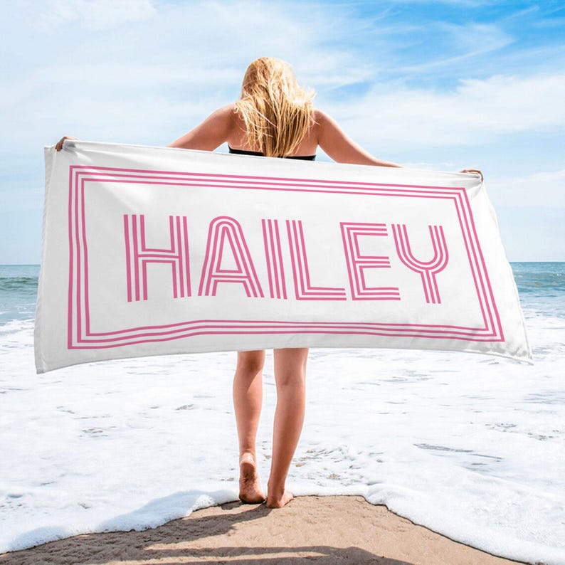 Puede incluir: Una toalla de playa blanca con el nombre "HAILEY" en rosa, enmarcada por l&iacute;neas rosas. La toalla es sostenida por una persona en una playa con el oc&eacute;ano y el cielo azul de fondo.